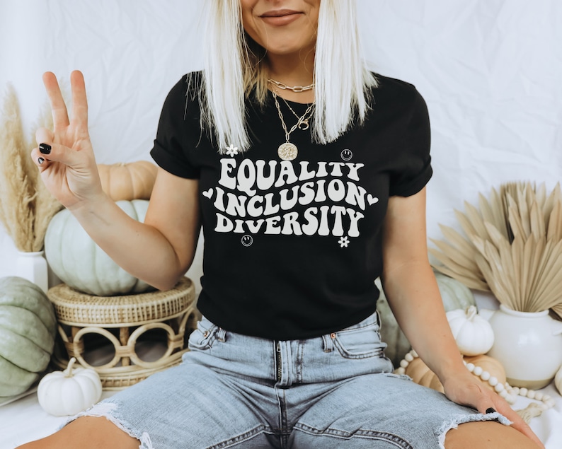 以下が含まれることがあります： レトロなフォントで「Equality Inclusion Diversity」と書かれた黒いTシャツを着た女性。彼女は、かぼちゃや秋の装飾品が置かれた白い表面に座っています。