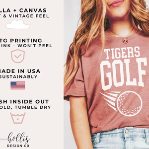 May include: A heather mauve t-shirt featuring the text "TIGERS GOLF" in white. A white golf ball with a white swoosh is positioned below the text. The shirt is crafted from Bella + Canvas fabric, known for its soft feel and vintage aesthetic. The shirt is printed using DTG printing, ensuring the ink won't peel. The shirt is sustainably made in the USA.