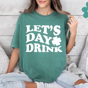 May include: A woman wearing a light green t-shirt with the text "Let's Day Drink" in white. The shirt features a white shamrock graphic.