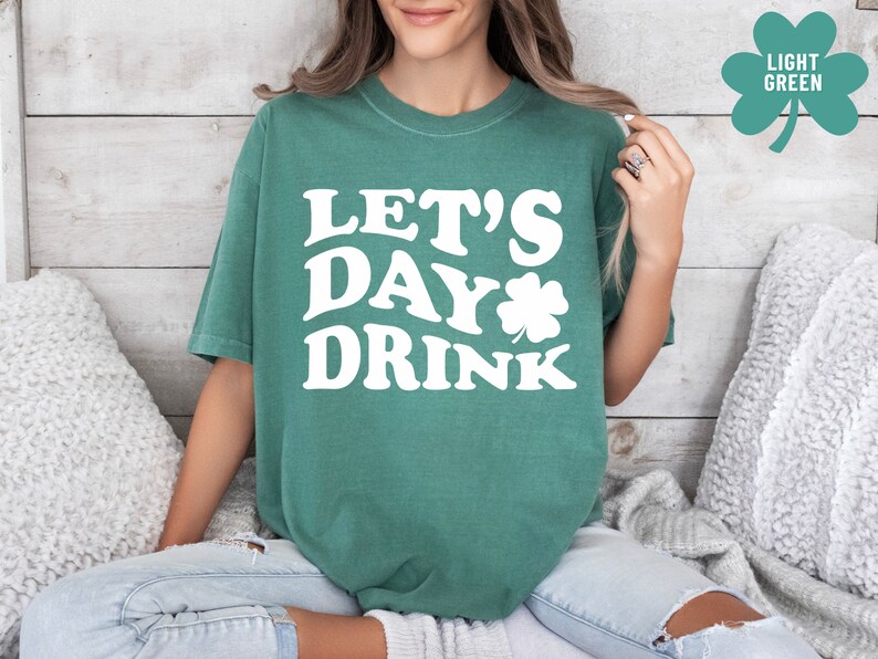 May include: A woman wearing a light green t-shirt with the text "Let's Day Drink" in white. The shirt features a white shamrock graphic.