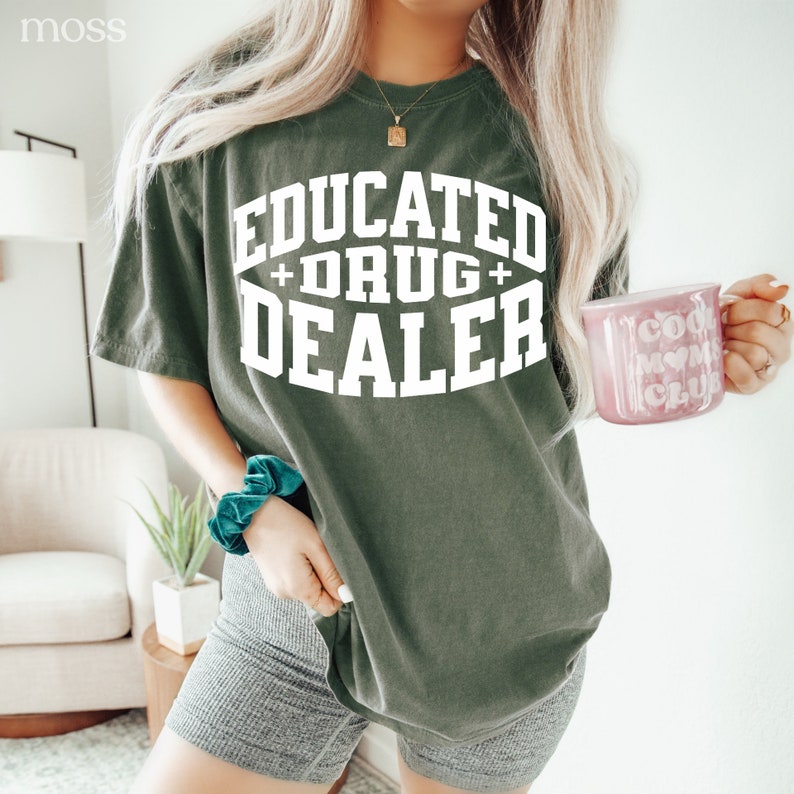 Op de afbeelding: Een groen T-shirt met de tekst "EDUCATED + DRUG + DEALER" in witte letters. Het shirt wordt gedragen door een persoon met lang blond haar. De persoon houdt een roze mok vast met de tekst "COOL MOM CLUB" in witte letters.