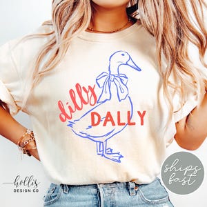 May include: Cream-coloured t-shirt with a blue and red duck graphic and the words "Dilly Dally". The shirt has short sleeves and is made of a soft material. The design is playful and whimsical.