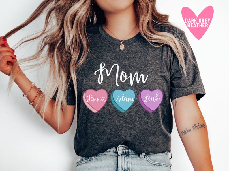 May include: A dark grey heather t-shirt with the word "mom" printed in white cursive. Three candy hearts with the names "Jenna", "Adam", and "Leah" are printed below the word "mom".