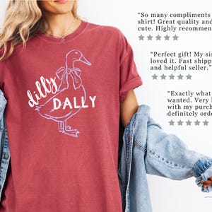 May include: Crimson t-shirt with a duck graphic and the words "dilly dally". The duck has a bow. The shirt is from Hollis Design Co. The image also includes customer reviews.