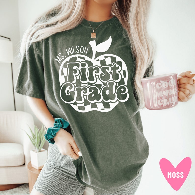 Puede incluir: Una camiseta verde oscuro con un gr&aacute;fico de manzana a cuadros blancos que dice "First Grade" y "Ms. Wilson" en texto blanco.