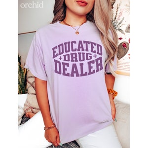 Op de afbeelding: Een lavendelkleurig T-shirt met de tekst "EDUCATED + DRUG + DEALER" in paars.