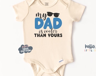 以下が含まれることがあります： クリーム色のベビーボディスーツ。黒と青で「my DAD is cooler THAN YOURS」と書かれています。「DAD」の文字は青色で、ボディスーツには「DAD」の文字の上に黒いサングラスが描かれています。ボディスーツには「SOFT 100% COTTON」の文字も書かれています。