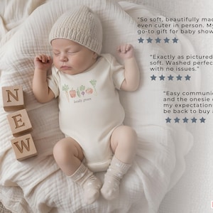 May include: A newborn baby wearing a beige knit cap and a cream-colored onesie with a "locally grown" vegetable design. Wooden blocks spell "NEW" next to the baby. The baby is lying on a soft, cream-colored blanket.