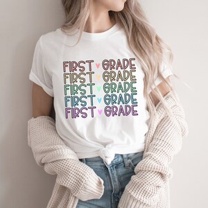 May include: White t-shirt with the text "FIRST ♥ GRADE" repeated five times in rainbow colors.