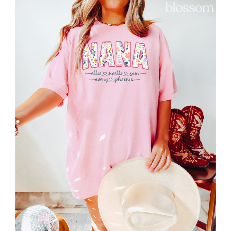 K&ouml;nnte beinhalten: Pinkes T-Shirt mit dem Wort "NANA" in Blumenbuchstaben. Das Shirt tr&auml;gt auch die Namen "Ellie, Noelle, Sam, Avery und Phoenix", die unter dem Wort "NANA" stehen.