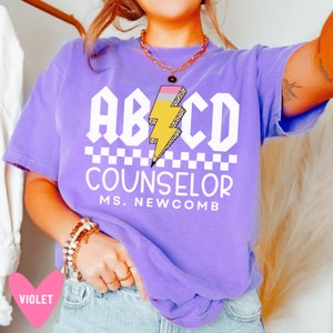 Pu&ograve; includere: Maglietta viola con una grafica bianca di un fulmine a forma di matita con il testo "ABCD COUNSELOR MS. NEWCOMB"
