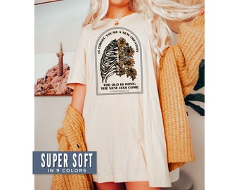 Op de afbeelding: Een wit T-shirt met een bloemenprint en de tekst "In Christus ben je een nieuwe schepping. Het oude is voorbij; het nieuwe is gekomen. 2 Korintiërs 5:17". Het shirt is superzacht en verkrijgbaar in 9 kleuren.