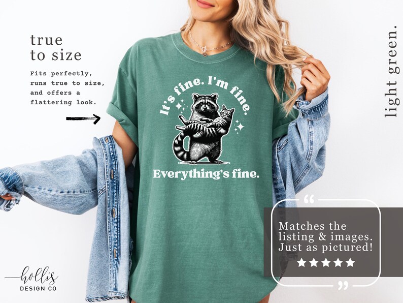 Puede incluir: Camiseta verde claro con un mapache sosteniendo un gato y el texto "It's fine. I'm fine. Everything's fine." La camiseta tiene un corte holgado y mangas enrolladas.