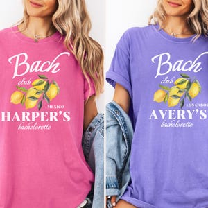 May include: Two t-shirts, one in a vibrant pink and the other in violet, each featuring a white graphic. The graphic includes the text "Bach club" above a lemon illustration and the text "Harper's bachelorette" and "Avery's bachelorette".
