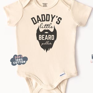 May include: Cream-colored baby bodysuit with black text that reads "DADDY'S little BEARD puller" and a beard graphic. The bodysuit has short sleeves and a snap closure. The words "SOFT 100% COTTON" are printed on a cloud-shaped graphic.