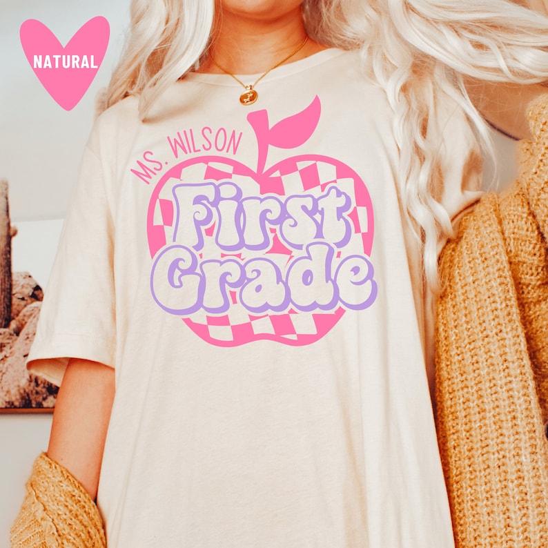 Puede incluir: Una camiseta blanca con un gr&aacute;fico de manzana rosa que dice "First Grade" en letras moradas. La manzana tiene un patr&oacute;n a cuadros y un tallo verde. El texto "Ms. Wilson" est&aacute; escrito sobre la manzana.