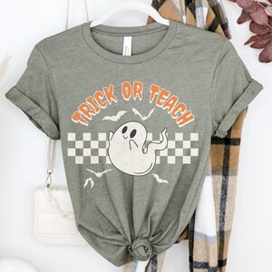 Op de afbeelding: Een groen T-shirt met een witte geestenafbeelding en de tekst "Trick or Teach" in oranje letters. Het shirt heeft een geruit patroon achter de geest.