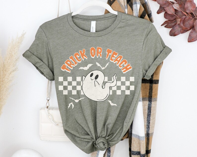 Op de afbeelding: Een groen T-shirt met een witte geestenafbeelding en de tekst "Trick or Teach" in oranje letters. Het shirt heeft een geruit patroon achter de geest.