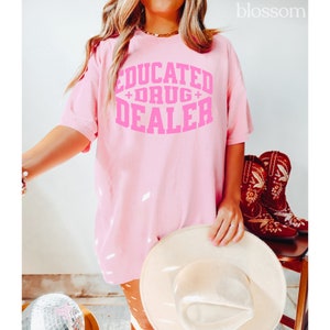 Op de afbeelding: Roze oversized T-shirt met de tekst "EDUCATED + DRUG + DEALER" in roze letters.