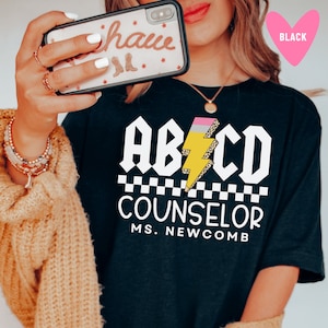 Pu&ograve; includere: Maglietta nera con una grafica bianca che dice "ABCD Counselor Ms. Newcomb". La grafica presenta un fulmine giallo con una gomma rosa e stampa leopardata.