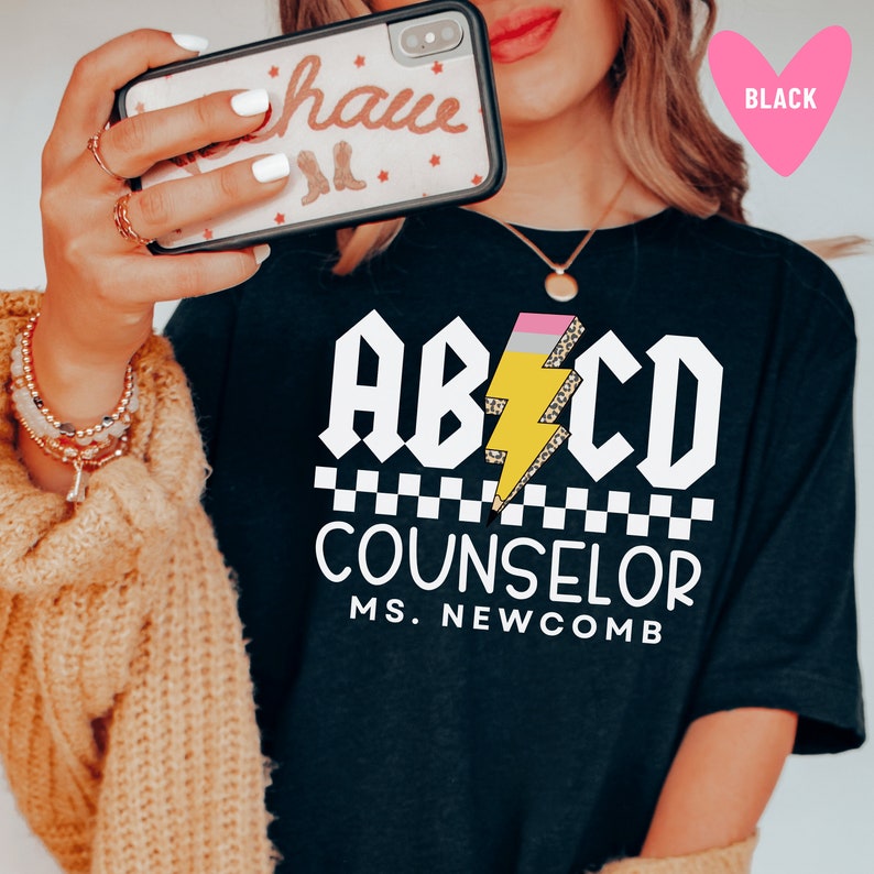 Pu&ograve; includere: Maglietta nera con una grafica bianca che dice "ABCD Counselor Ms. Newcomb". La grafica presenta un fulmine giallo con una gomma rosa e stampa leopardata.