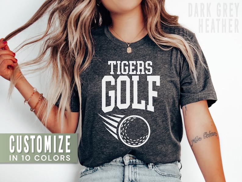 May include: A dark grey heather t-shirt with the text "TIGERS GOLF" in white. A white golf ball with a white stripe is flying behind the text. The shirt is available in 10 colours.