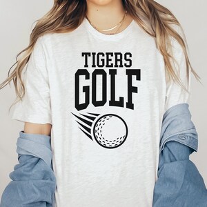 May include: White t-shirt with black text that reads "TIGERS GOLF" and a black and white golf ball graphic.