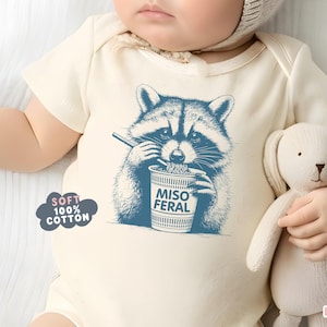 May include: Cream-colored baby bodysuit with a blue graphic of a raccoon eating noodles from a cup that says "Miso Feral." The bodysuit also has the text "Soft 100% Cotton." A baby is wearing the bodysuit and holding a stuffed animal.