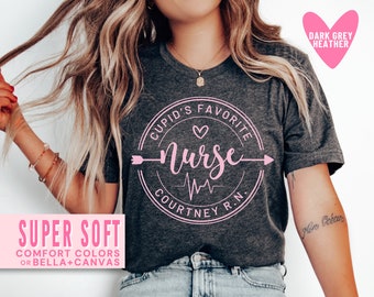 May include: A dark grey heather t-shirt with a pink circle graphic that says "Cupid's Favorite Nurse" with a heart, arrow, and EKG line. The text "Courtney R.N." is also included in the graphic.