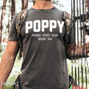 May include: A dark gray t-shirt with the word "POPPY" printed in white block letters. Below the word "POPPY" are the names "PHOENIX - RYDER - OLIVIA MASON - AVA" printed in white.