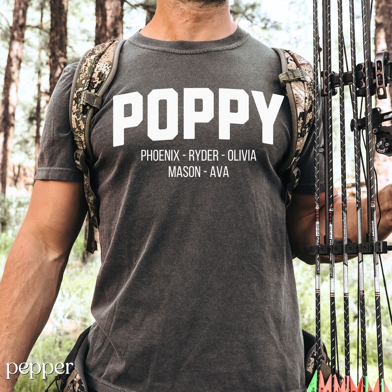 May include: A dark gray t-shirt with the word "POPPY" printed in white block letters. Below the word "POPPY" are the names "PHOENIX - RYDER - OLIVIA MASON - AVA" printed in white.