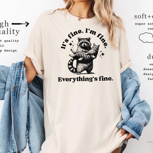 Puede incluir: Camiseta color marfil con un mapache abrazando a un gato, con el texto "It's fine. I'm fine. Everything's fine." La camiseta tambi&eacute;n tiene texto que dice "alta calidad" y "duradero".