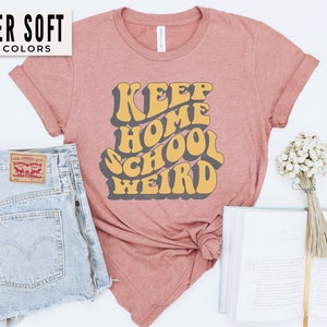 May include: A pink t-shirt with the text "Keep Home School Weird" in a retro font. The text is in yellow and gray.