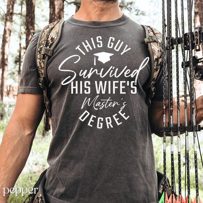 Peut inclure: Un t-shirt gris avec un texte blanc qui dit "This Guy Survived His Wife's Master's Degree". Le texte est dans un cercle avec une ic&ocirc;ne de chapeau de graduation au-dessus du mot "Survived".