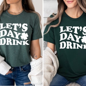 May include: A green t-shirt with white text that says "Let's Day Drink" and a shamrock graphic. The shirt is shown on a model wearing blue jeans and a white cardigan.