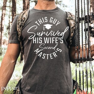 May include: A grey t-shirt with white text that reads "This Guy Survived His Wife's Second Master's". The text is in a circle with a graduation cap icon above the word "Survived".