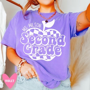 May include: Purple t-shirt with a white checkered apple design and the text "Ms. Wilson Second Grade".