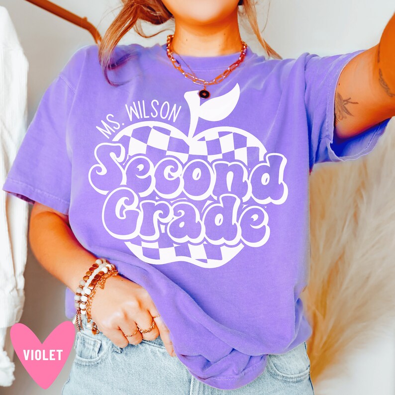 May include: Purple t-shirt with a white checkered apple design and the text "Ms. Wilson Second Grade".