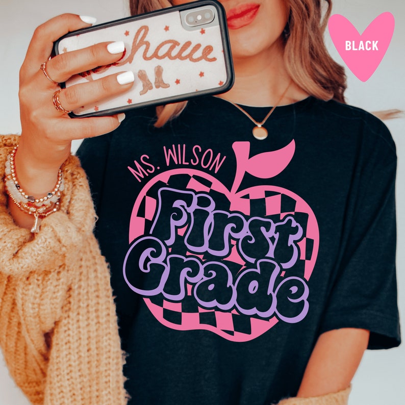Puede incluir: Camiseta negra con un dise&ntilde;o de manzana rosa que dice "Ms. Wilson First Grade" en letras moradas y negras.