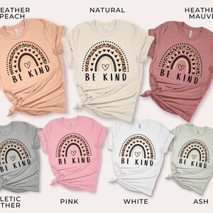 May include: Seven t-shirts in different colors featuring a rainbow with a heart and the text "Be Kind". The rainbow is decorated with leopard print and hearts. The colors of the t-shirts are heather peach, natural, heather mauve, athletic heather, pink, white, and ash.