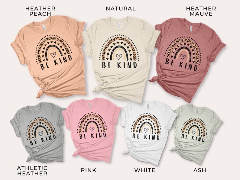 May include: Seven t-shirts in different colors featuring a rainbow with a heart and the text "Be Kind". The rainbow is decorated with leopard print and hearts. The colors of the t-shirts are heather peach, natural, heather mauve, athletic heather, pink, white, and ash.