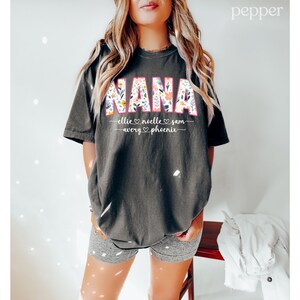 K&ouml;nnte beinhalten: Ein schwarzes T-Shirt mit dem Wort "NANA" in gro&szlig;en, bunten Buchstaben mit Blumenmustern. Der Text "ellie &hearts; noelle &hearts; sam - avery &hearts; phoenix" ist unter dem Wort "NANA" gedruckt.
