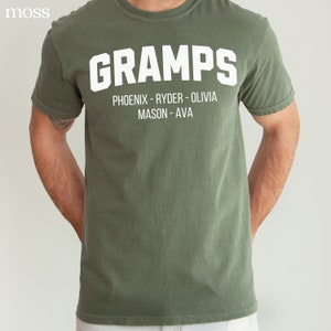 May include: A green t-shirt with the word "GRAMPS" in white letters. Below the word is the text "PHOENIX - RYDER - OLIVIA MASON - AVA" in white letters.