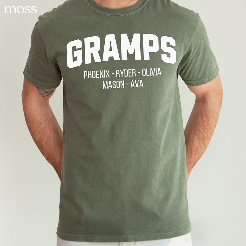 May include: A green t-shirt with the word "GRAMPS" in white letters. Below the word is the text "PHOENIX - RYDER - OLIVIA MASON - AVA" in white letters.
