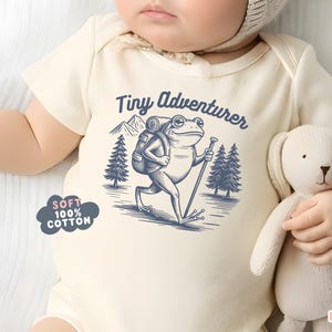 May include: Cream-colored baby onesie with a blue illustration of a frog adventurer, mountains, and trees. Text reads "Tiny Adventurer." A cloud-shaped graphic states "Soft 100% Cotton."