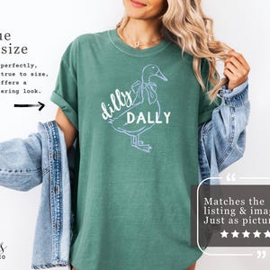 May include: A light green t-shirt featuring a white outline of a duck and the words "dilly dally". The shirt has rolled-up sleeves and is made of a soft material. The text "true to size" is also visible.