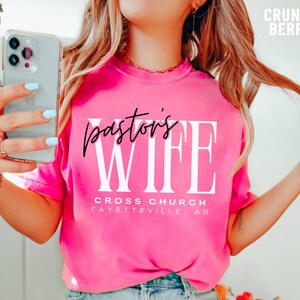 May include: Pink t-shirt with the text "pastor's wife" in black and white lettering. The text "Cross Church Fayetteville, AR" is printed below the main text.