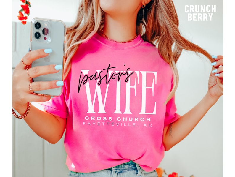 May include: Pink t-shirt with the text "pastor's wife" in black and white lettering. The text "Cross Church Fayetteville, AR" is printed below the main text.