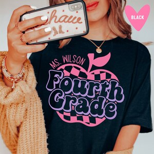 K&ouml;nnte beinhalten: Schwarzes T-Shirt mit einem pinkfarbenen und schwarzen karierten Apfel-Motiv und dem Text "Ms. Wilson Fourth Grade" in lila und schwarzen Buchstaben.