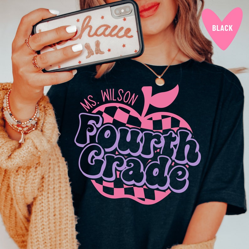 K&ouml;nnte beinhalten: Schwarzes T-Shirt mit einem pinkfarbenen und schwarzen karierten Apfel-Motiv und dem Text "Ms. Wilson Fourth Grade" in lila und schwarzen Buchstaben.